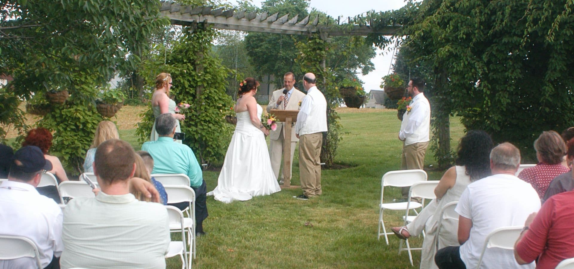 A couple exchanges vows during an outdoor wedding ceremony under a trellis, surrounded by guests.