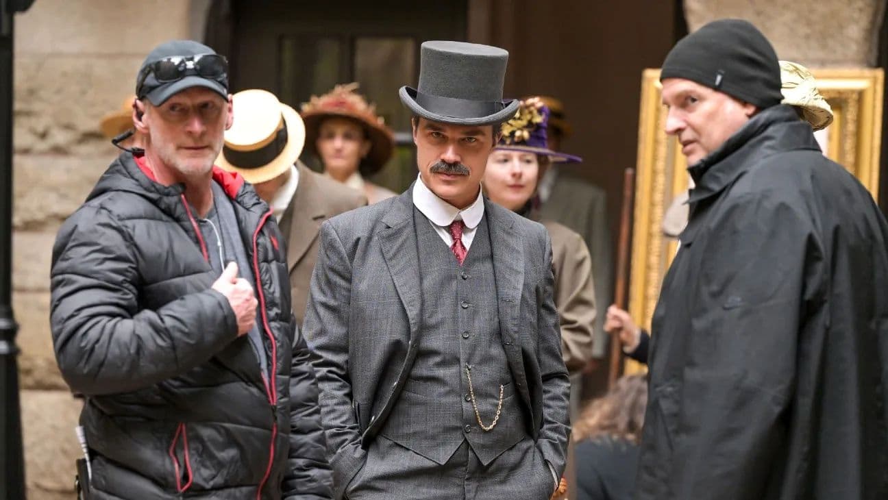 A group of people, including a man in a top hat and formal attire, are on a movie set surrounded by crew members.