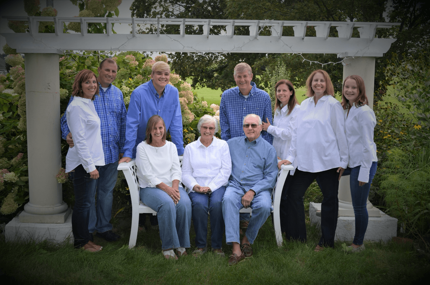 A family group portrait outdoors, smiling and dressed in white and blue attire, gathered under a trellis surrounded by greenery.