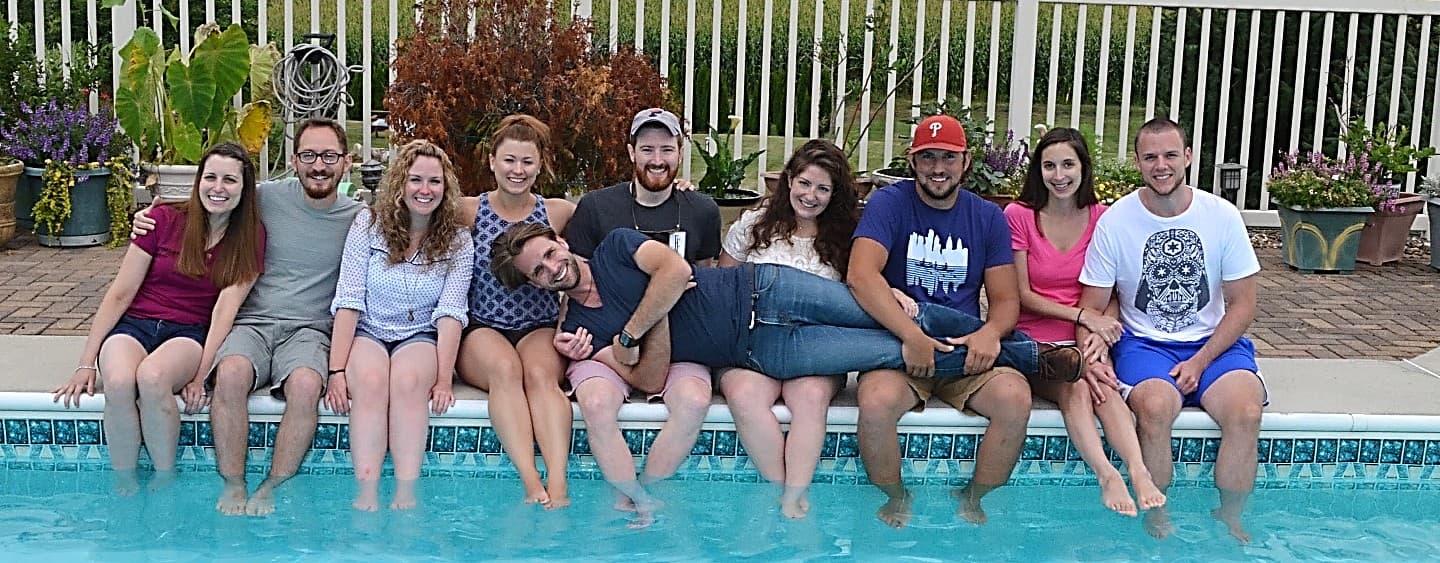 A group of ten friends sitting side by side on the edge of a pool, smiling and enjoying a sunny day.