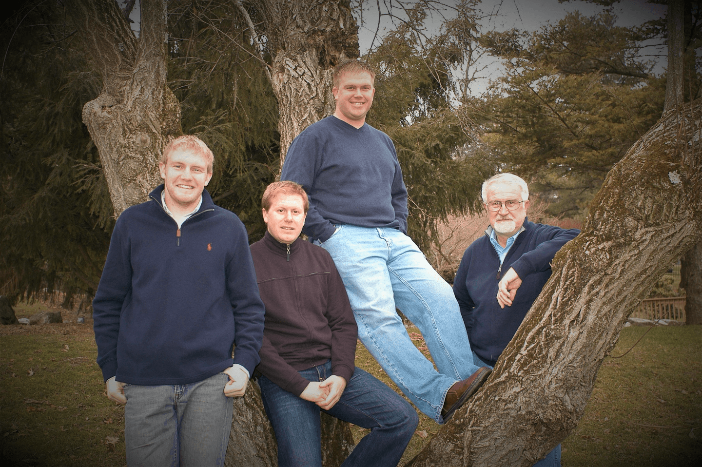 Four men stand together on a tree in a park, each wearing casual sweaters.