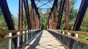 A rustic metal bridge spans a tree-lined path under a clear blue sky.