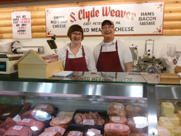 Two smiling employees stand behind a deli counter filled with various meats and cheeses at S. Clyde Weaver.