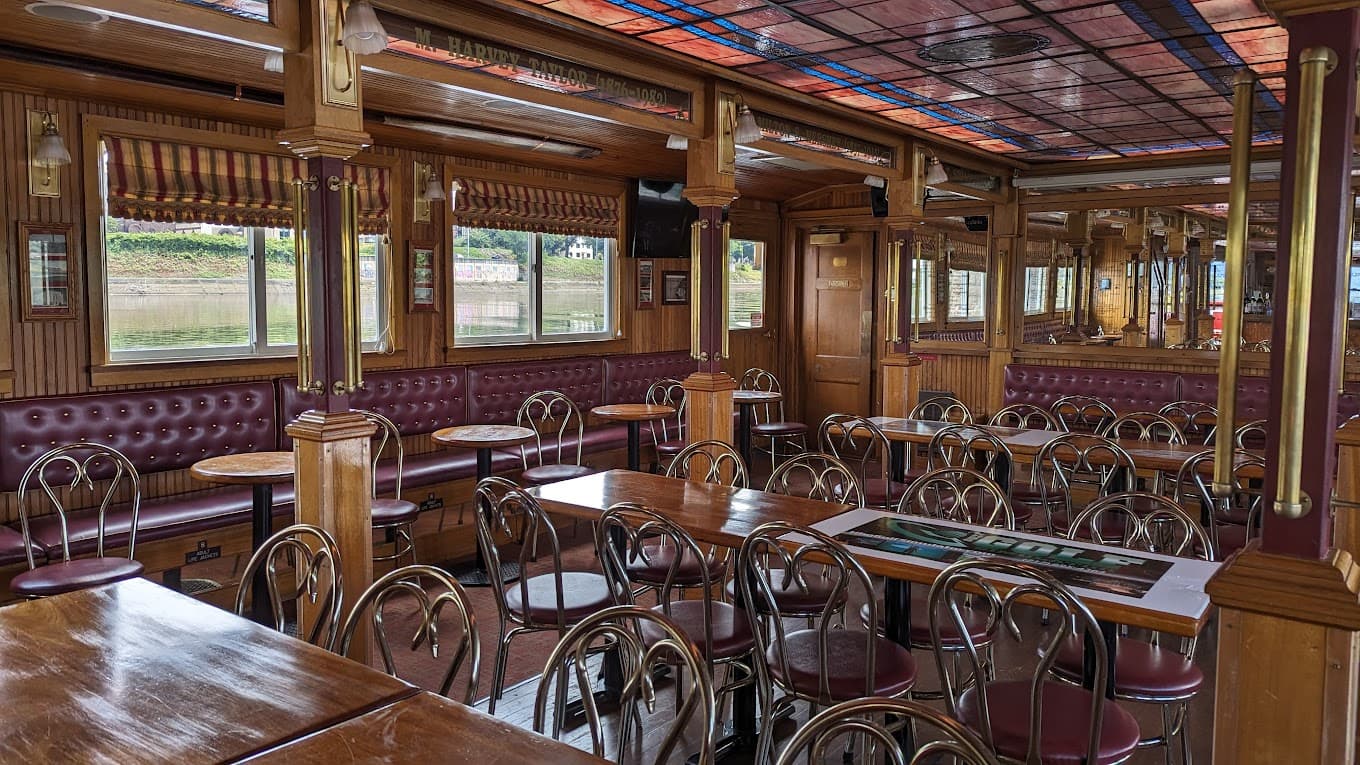 Interior of a cozy restaurant with wooden decor, tables, and chairs, featuring large windows.