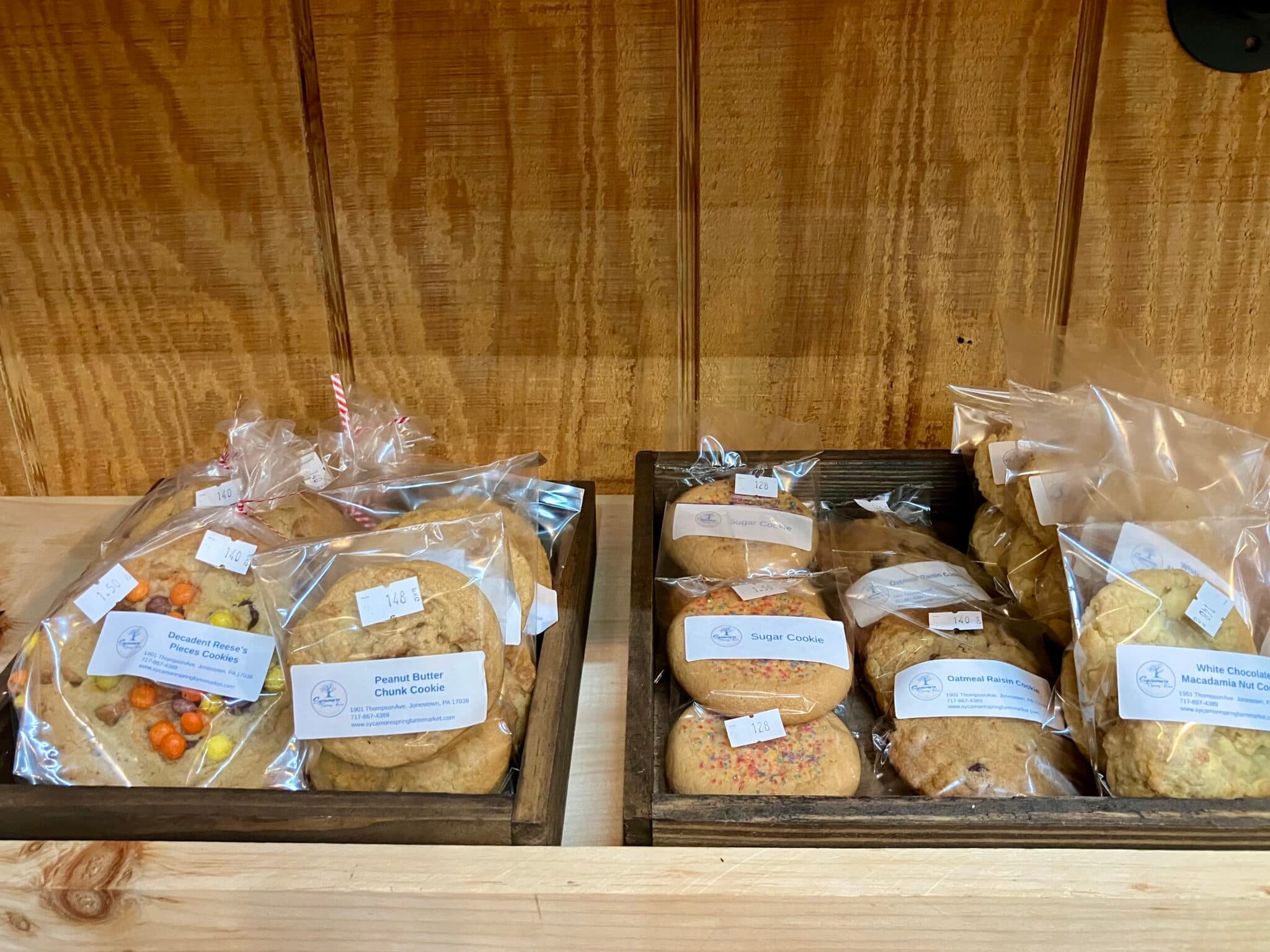 A wooden shelf displays various types of packaged cookies, including peanut butter, sugar, and white chocolate macadamia.