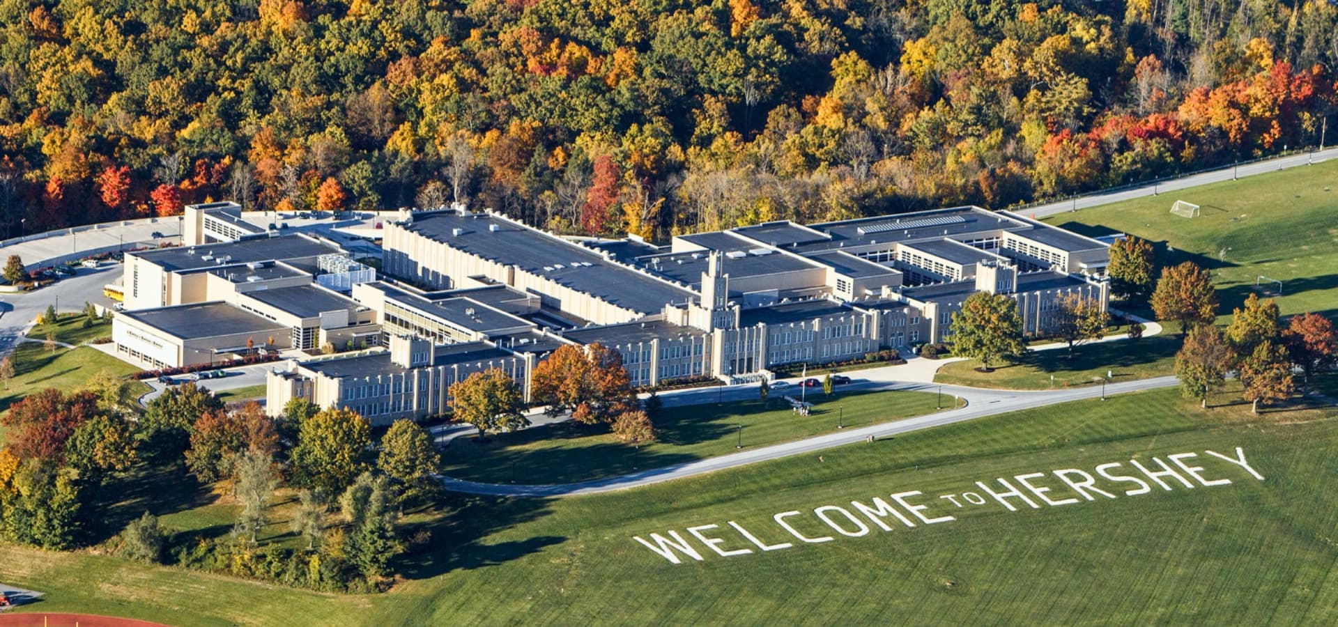 Aerial view of a large building surrounded by colorful autumn foliage, with "WELCOME TO HERSHEY" spelled out on the grass below.