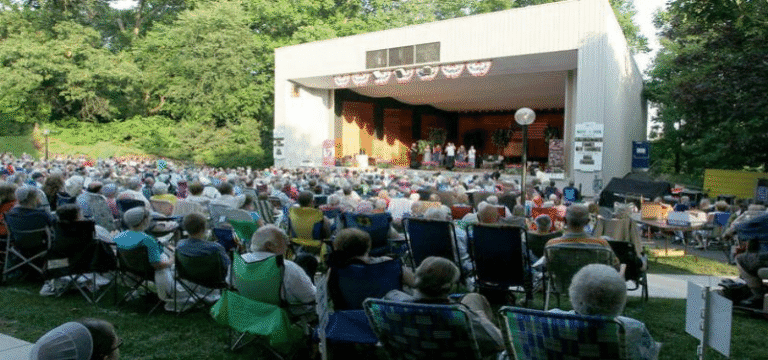A large crowd of people sits in lawn chairs facing a stage during an outdoor event.