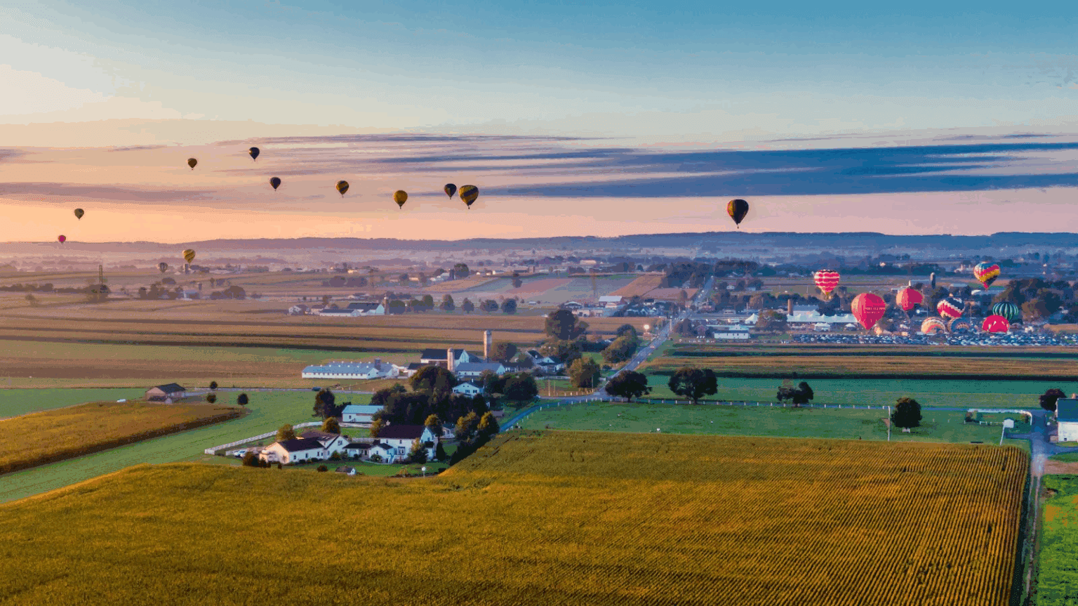 Aerial view of a countryside landscape with colorful hot air balloons floating above fields and a small town at sunrise.