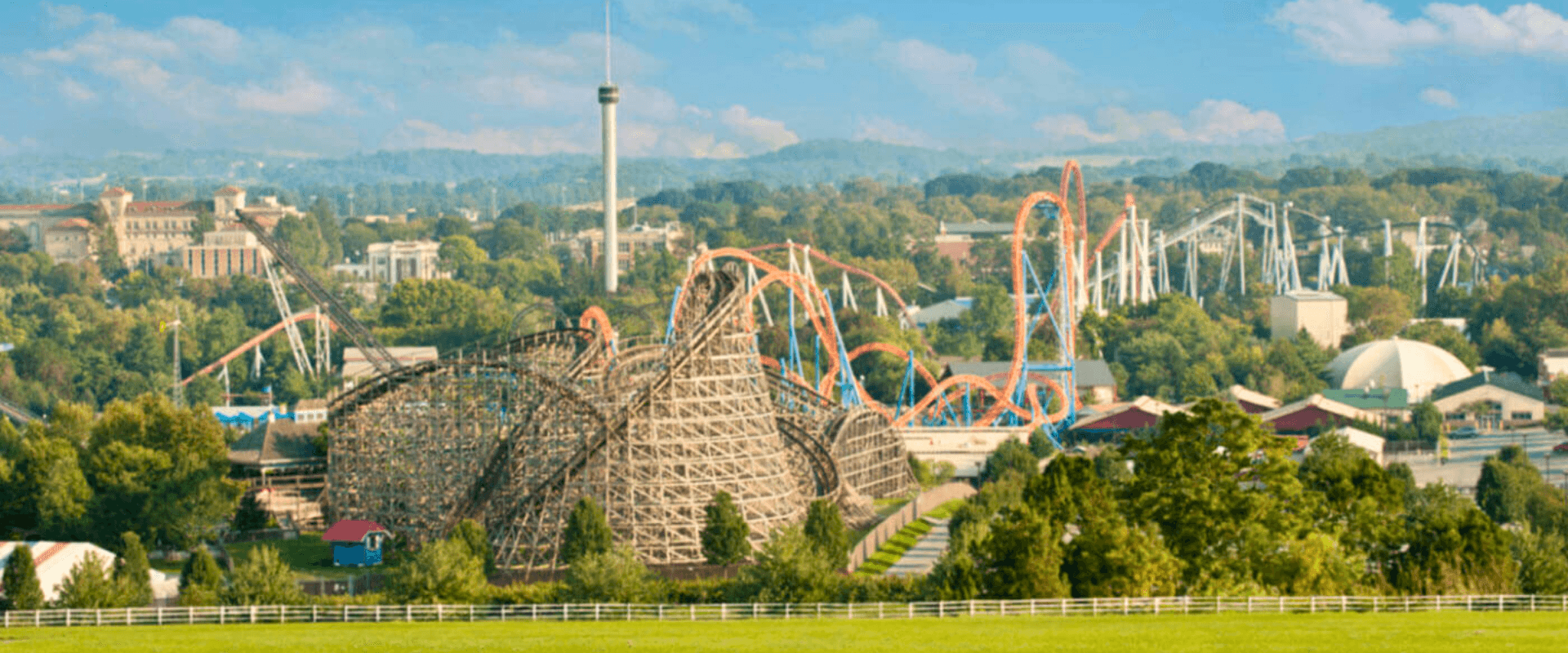 A panoramic view of a theme park featuring a large wooden roller coaster and various colorful rides amidst lush greenery.