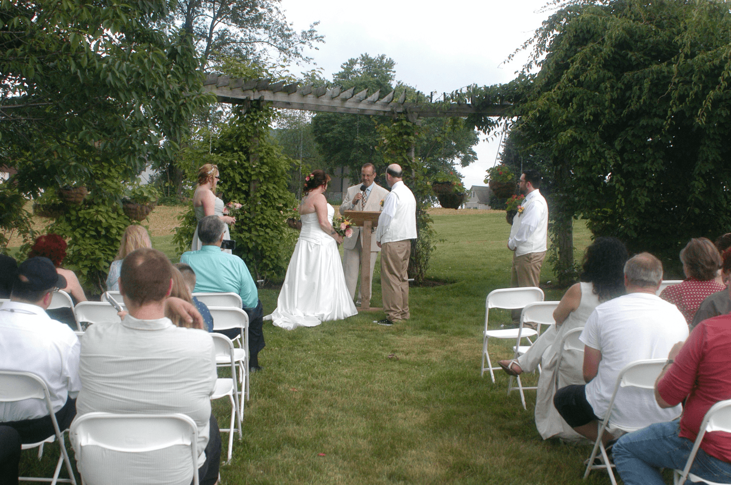 A bride and groom exchange vows under a trellis as guests look on during an outdoor wedding ceremony.