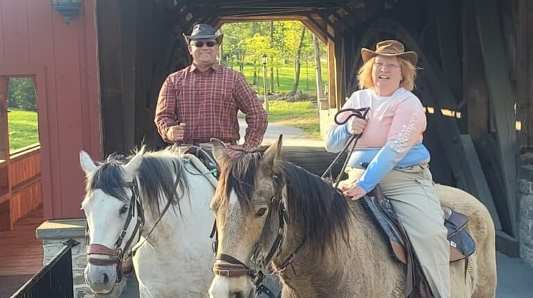 Two people on horseback smile while standing under a covered bridge in a green outdoor setting.