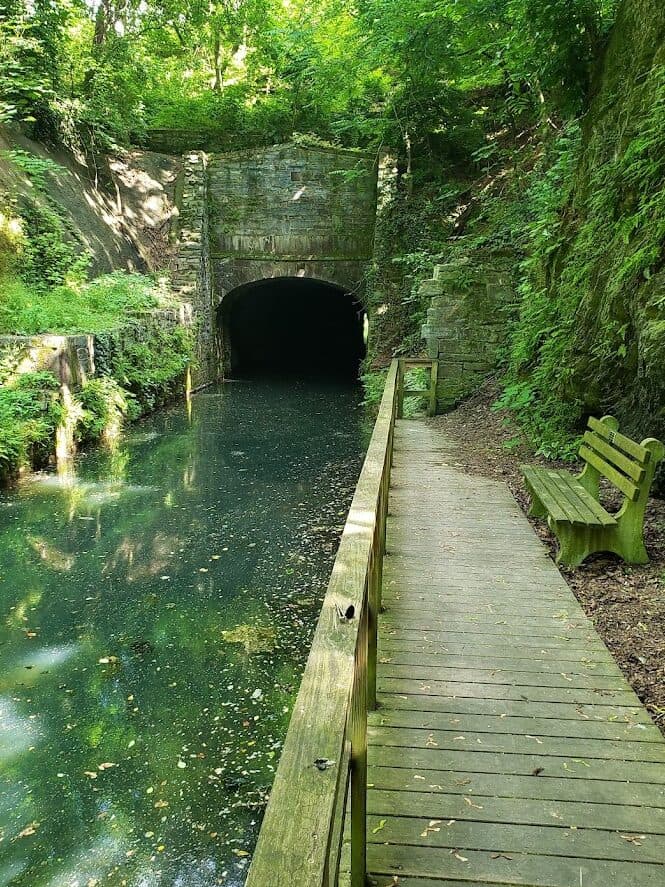 A serene pathway along a canal leading to a dark stone tunnel, surrounded by lush greenery.