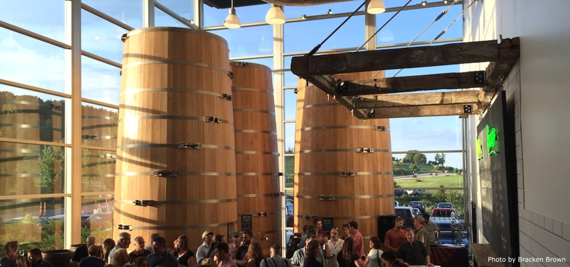 Interior view of a brewery with large wooden fermentation tanks and a crowd of people.