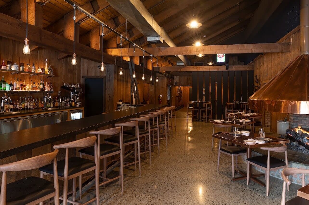 A rustic restaurant interior features a long wooden bar, high stools, and cozy dining tables near a fireplace.