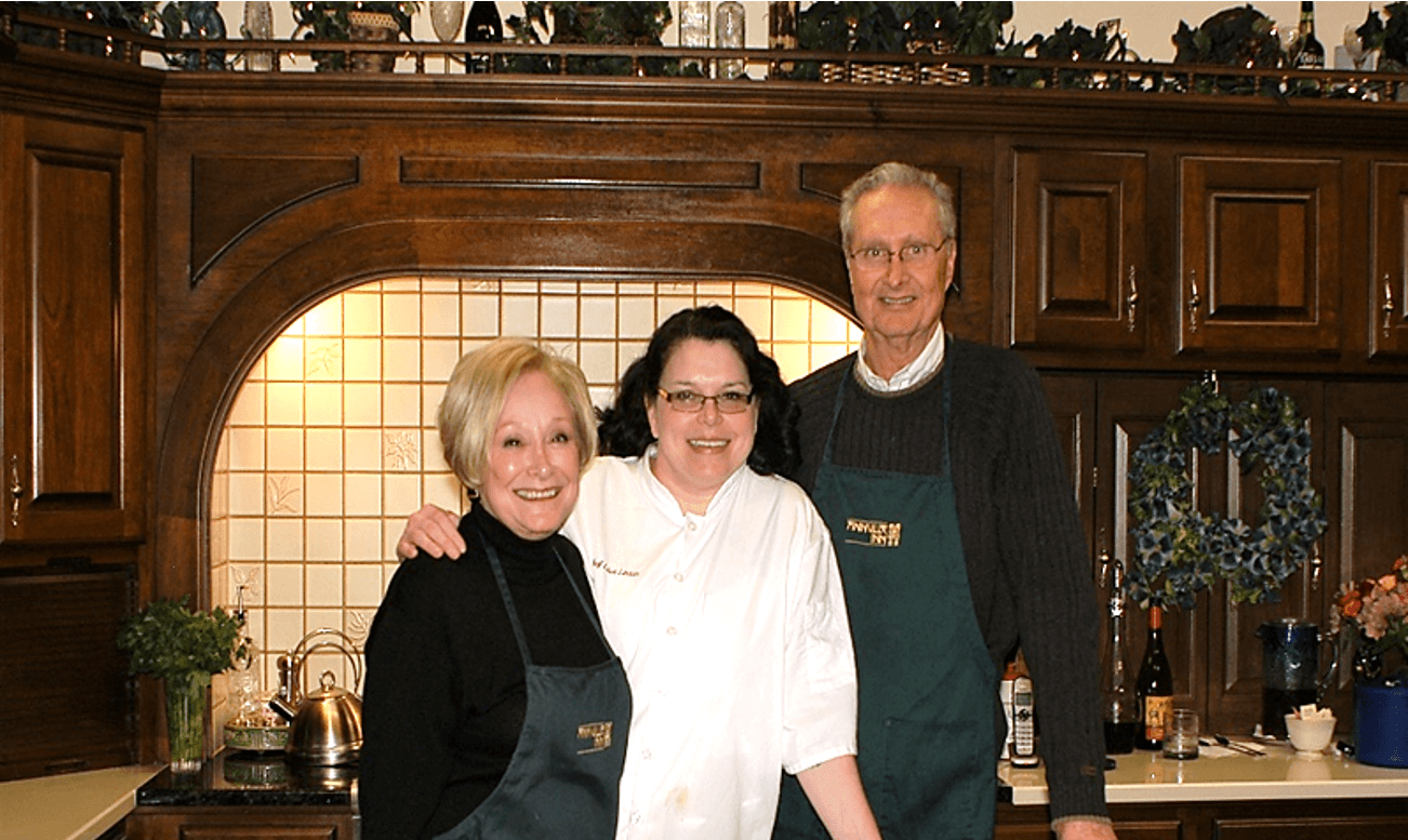 Three smiling individuals stand together in a kitchen, wearing aprons and posing for the photo.