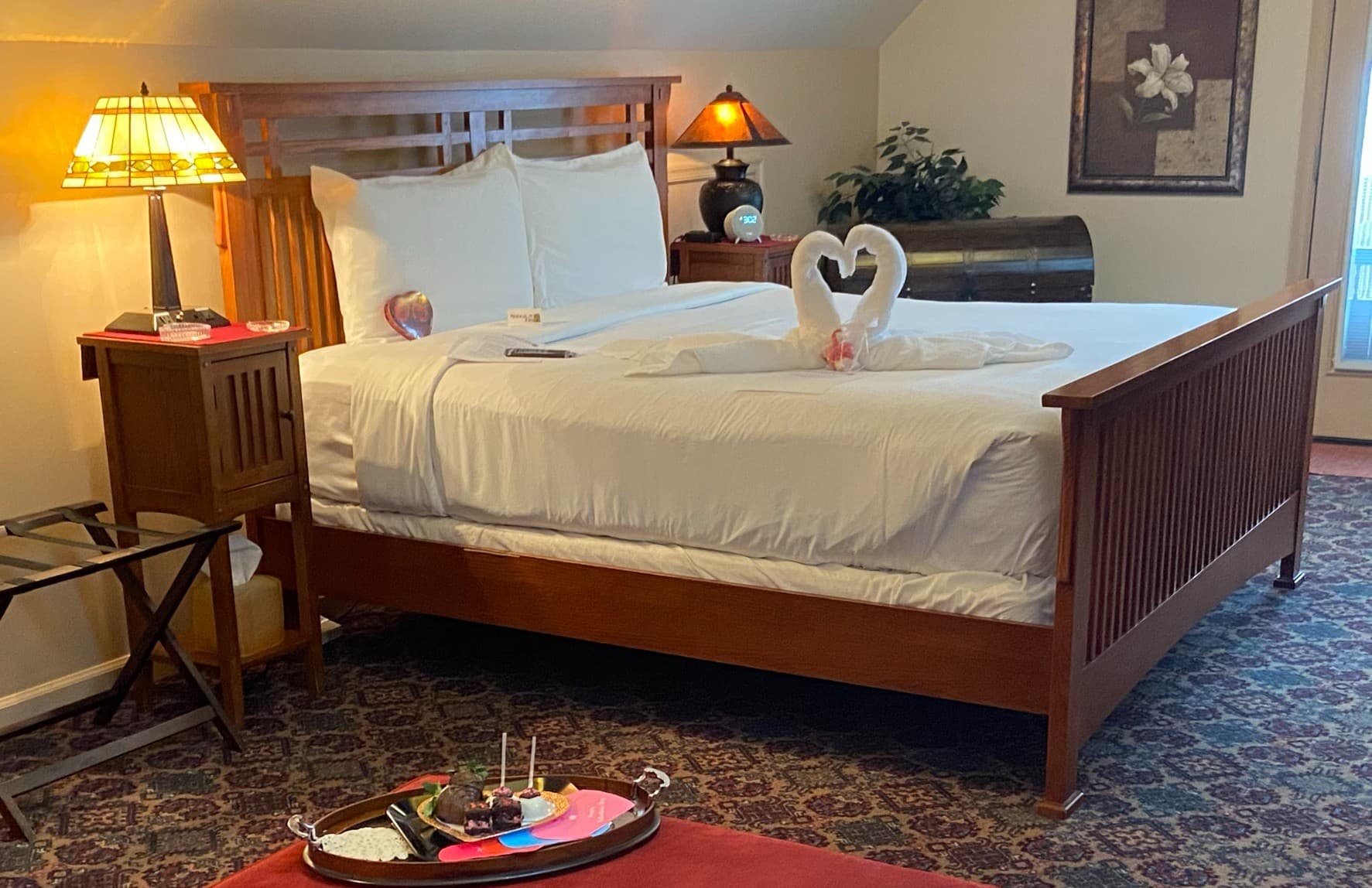 A cozy bedroom featuring a large bed with decorative swans made of towels, soft lighting, and a tray of snacks.