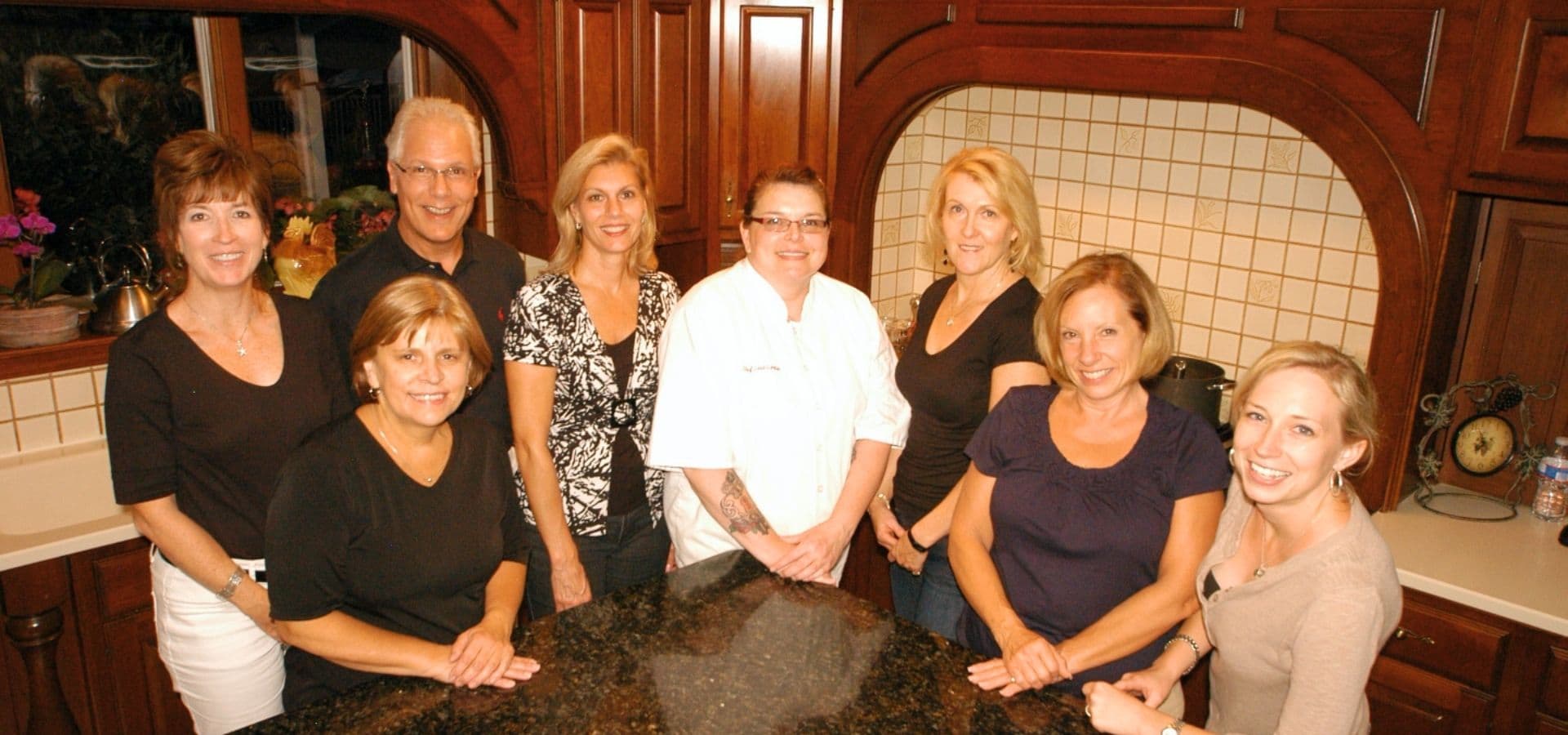 A group of eight adults stands around a kitchen island, smiling for the camera.