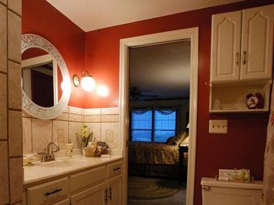 A cozy bathroom with red walls, a mirror, and a doorway leading into a softly lit bedroom.