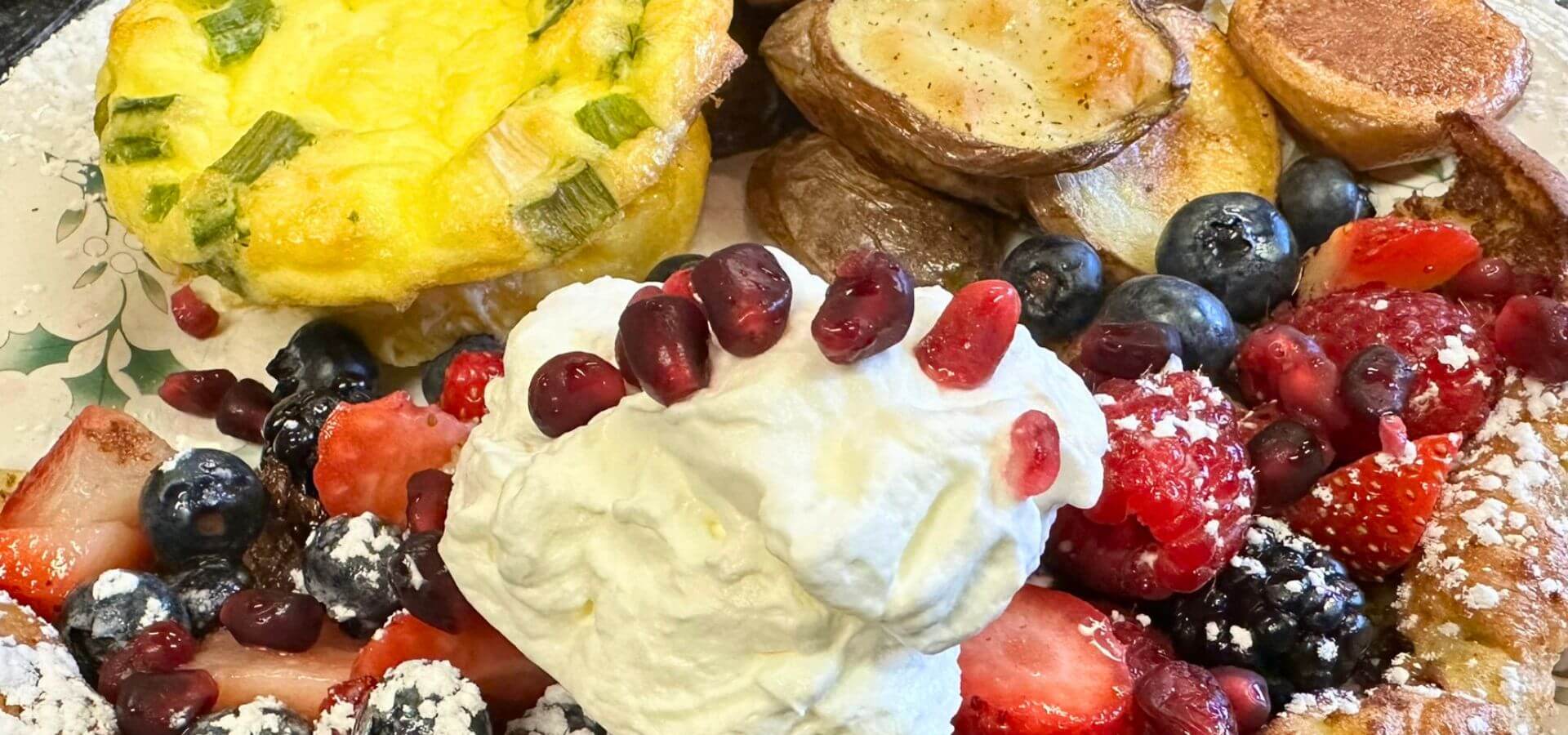 A colorful breakfast plate featuring an egg frittata, roasted potatoes, and a variety of fresh berries topped with whipped cream.