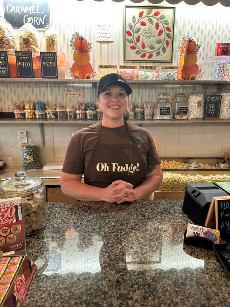 A smiling woman in an apron stands behind a candy shop counter filled with treats.
