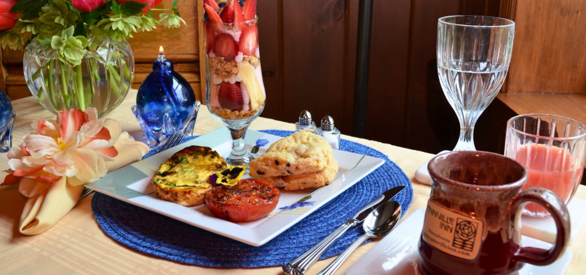 A beautifully arranged breakfast plate featuring a frittata, roasted tomato, scone, and a parfait, set on a table with decorative flowers and beverages.