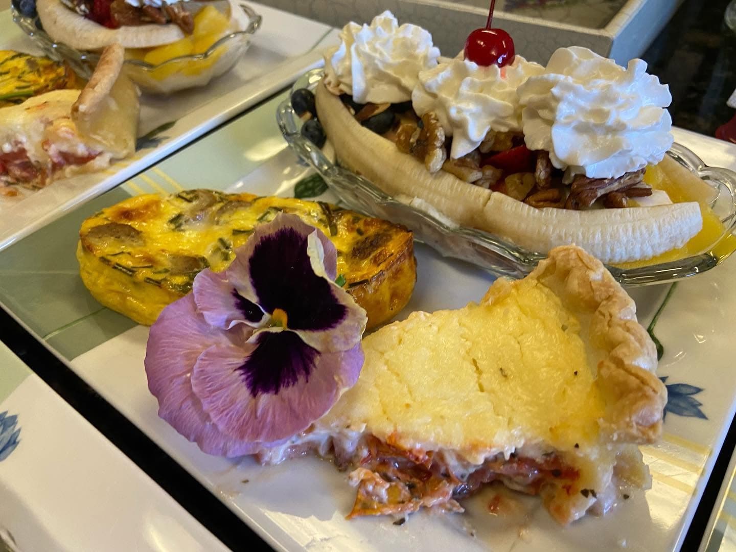 A plate featuring a colorful quiche, a slice of savory pie, and a banana split topped with whipped cream and a cherry, accompanied by a purple flower.