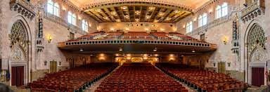 A panoramic view of a grand theater interior featuring rows of red seats and ornate architectural details.