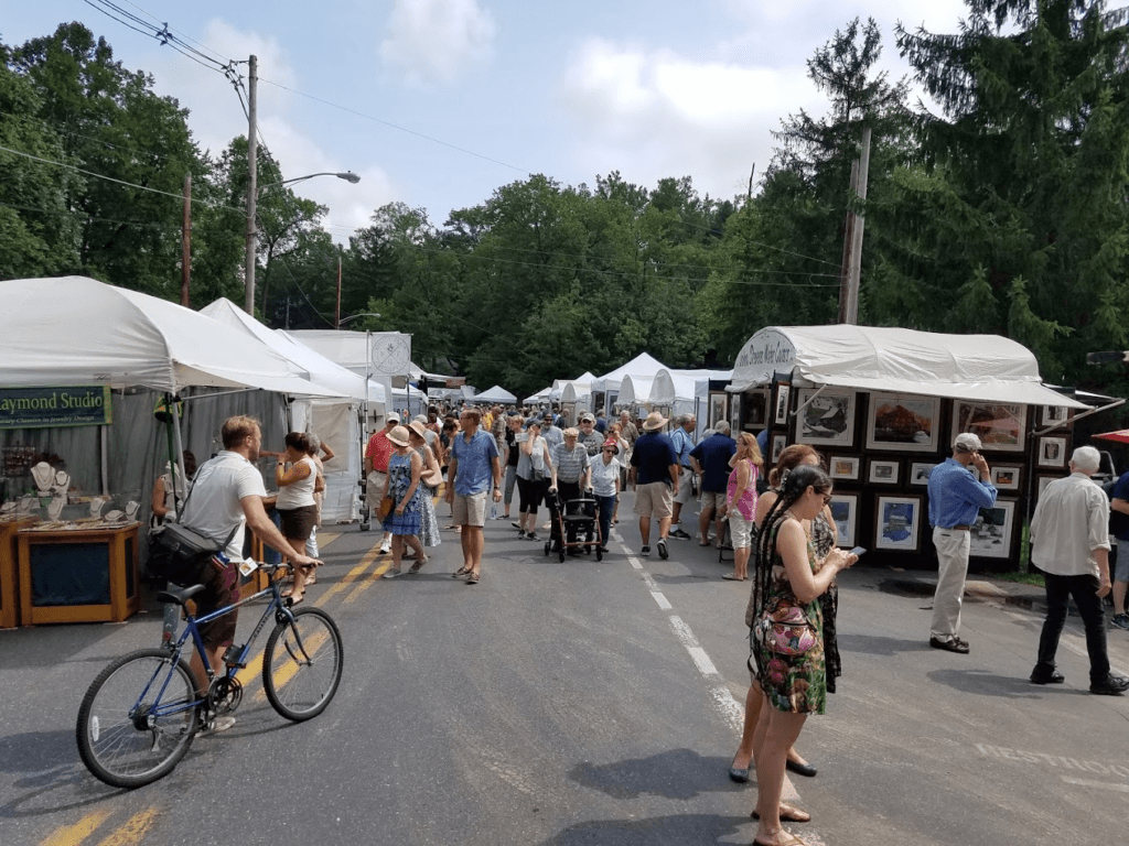 A busy street fair with art and craft vendors, crowded with people enjoying the event.