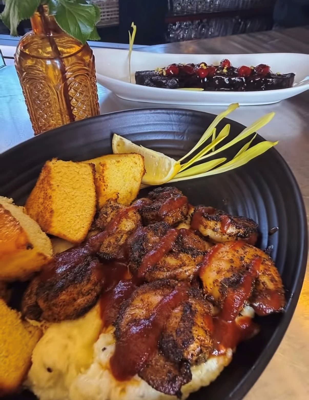 A black bowl filled with shrimp drizzled in sauce, cornbread, and a slice of lemon, with a dessert in the background.