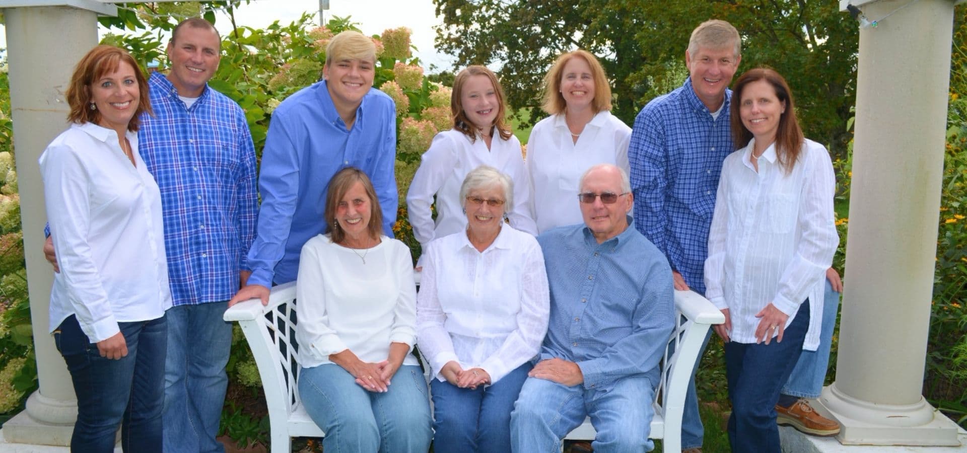 A group of ten people poses together outdoors, dressed in varying shades of blue and white, sitting and standing around a white bench in a garden setting.