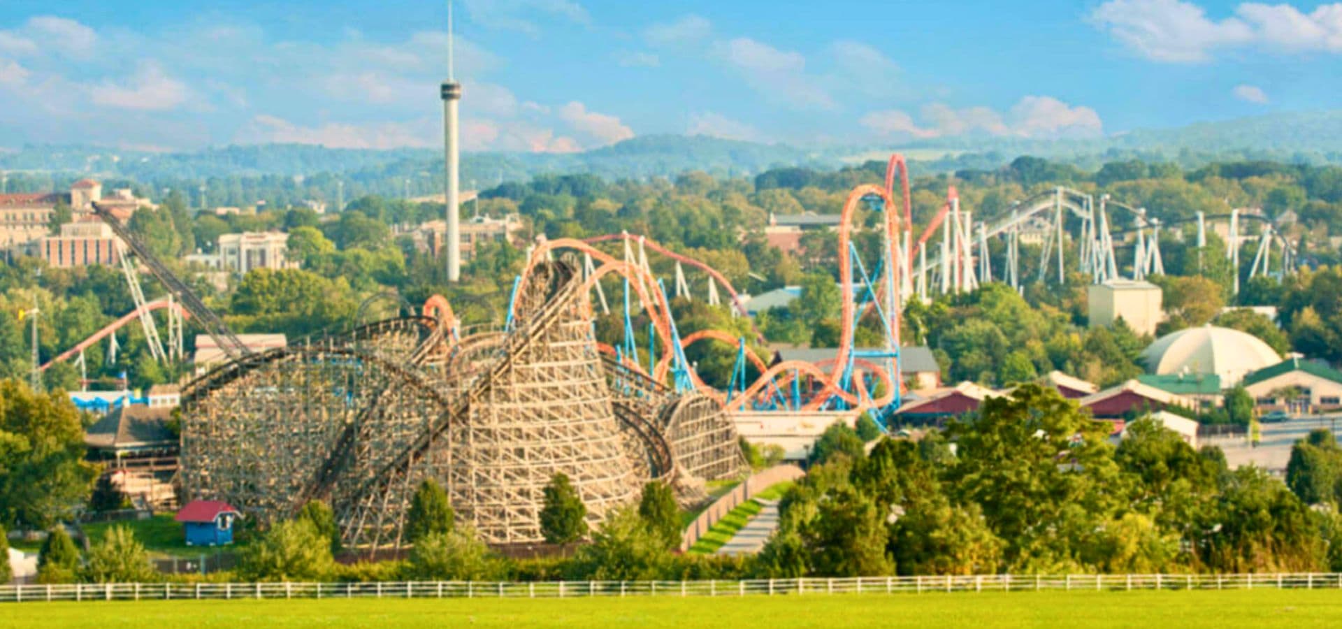 A panoramic view of an amusement park featuring a wooden roller coaster and colorful rides amid green trees and blue skies.