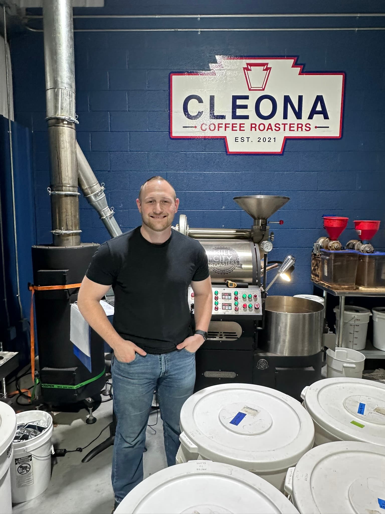 A smiling man stands in front of a coffee roasting machine in a roastery.