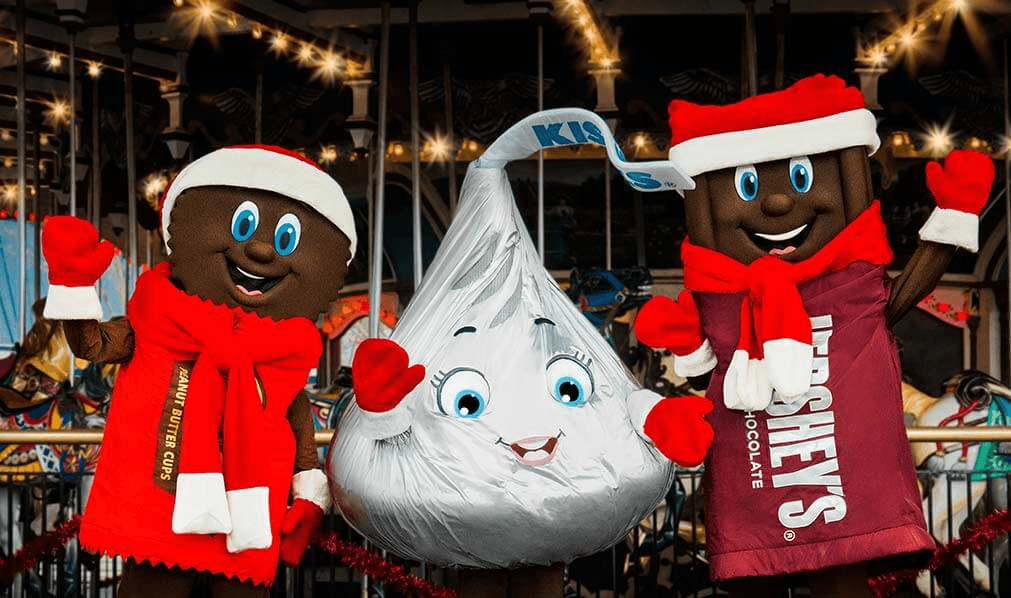 Three cartoonish chocolate mascots, including a Hershey's Kiss, pose festively wearing Santa hats.