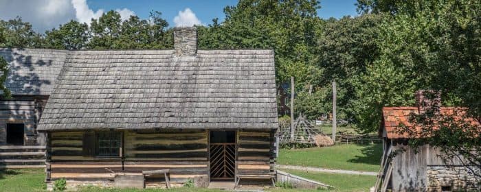 A rustic log cabin with a stone chimney surrounded by greenery.