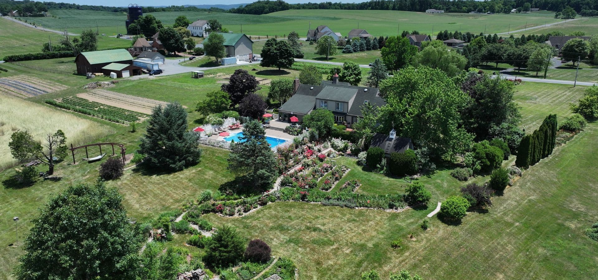 Aerial view of a lush garden with a swimming pool surrounded by a residential area and fields.