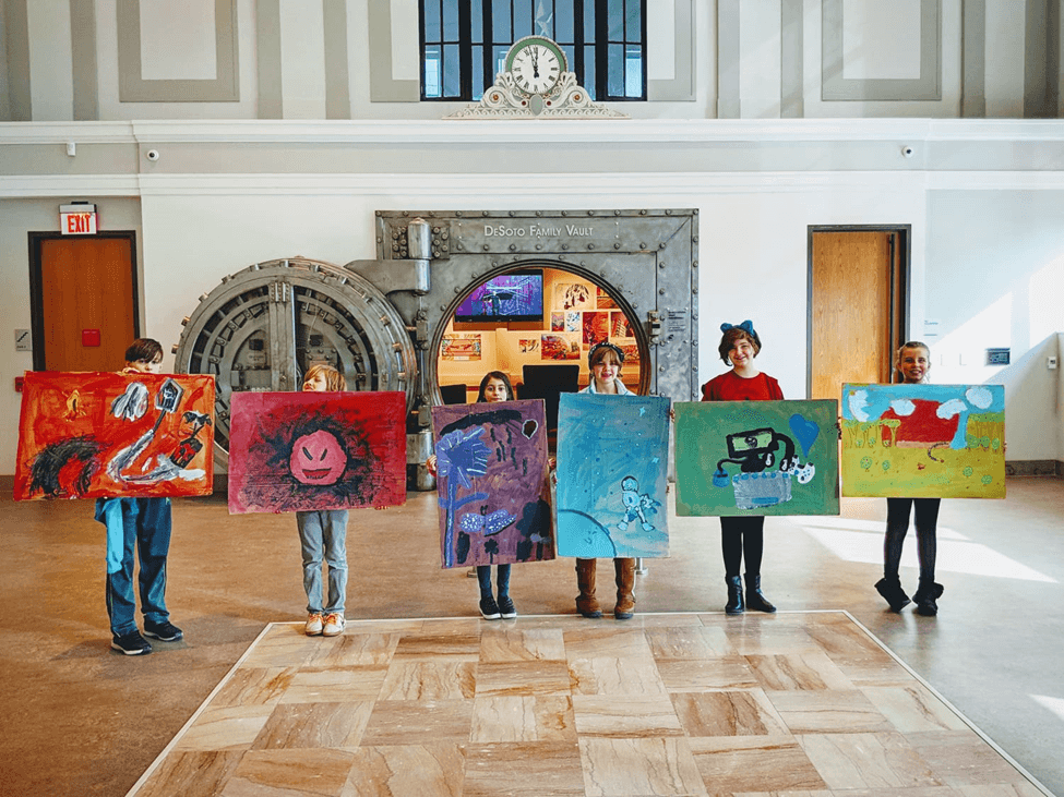 A group of six children stands in a brightly lit room, each holding a colorful piece of art in front of a large vault door.