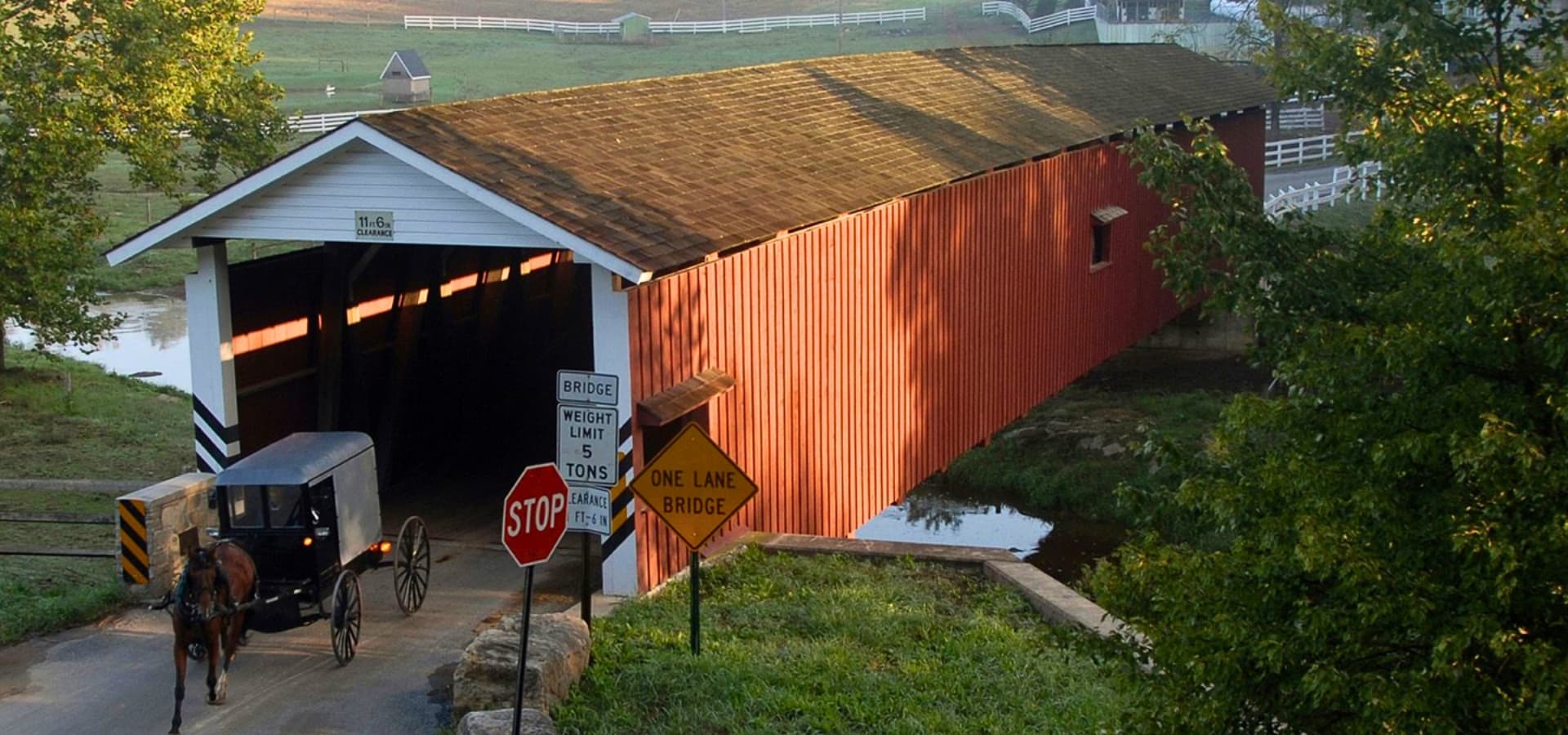 A horse-drawn buggy approaches a red covered bridge with road signs indicating traffic restrictions.