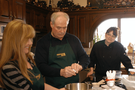 Three people, two women and one man, are cooking in a warmly lit kitchen, with one woman smiling and another observing as the man prepares ingredients.