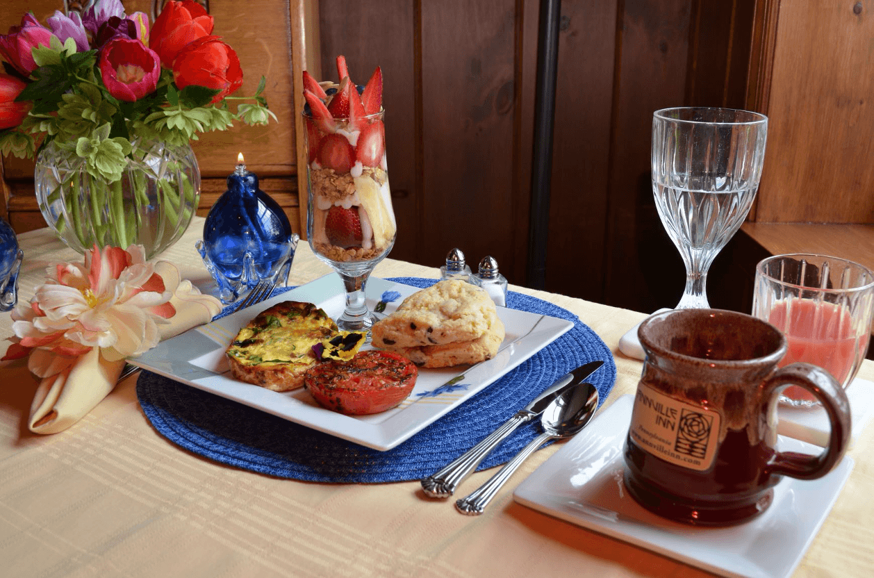 A beautifully arranged breakfast table featuring a vegetable frittata, a fruit parfait, a scone, and drinks, with colorful flowers as a centerpiece.