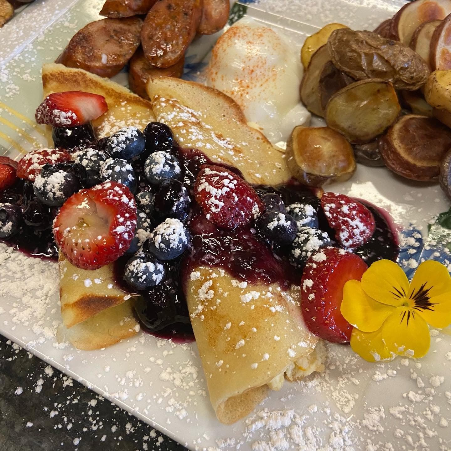 A plate featuring crepes topped with mixed berries and sauce, garnished with powdered sugar and a flower, alongside sausage, a poached egg, and roasted potatoes.