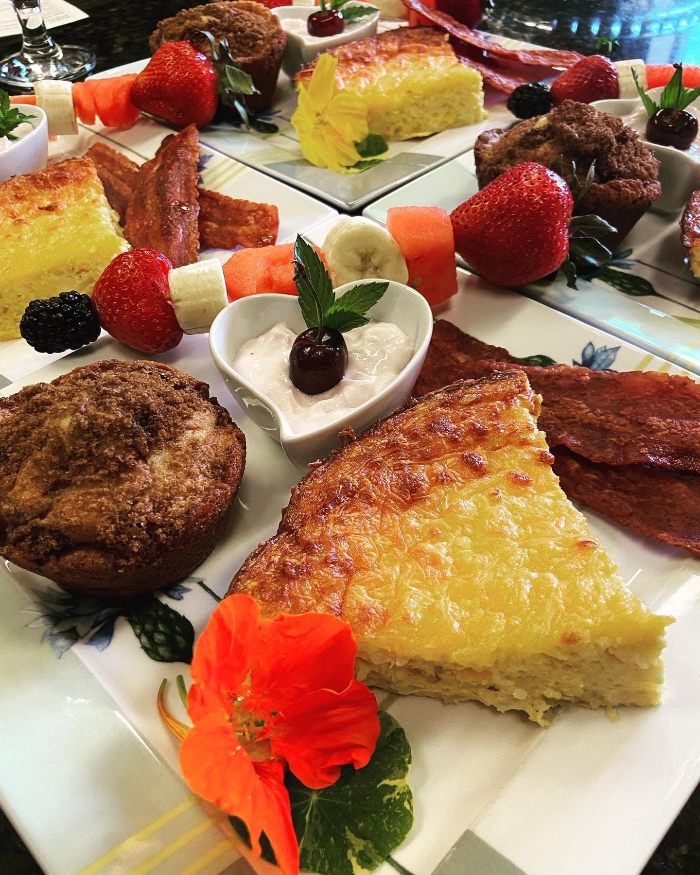 A colorful breakfast spread featuring quiche, muffins, fruit, bacon, and yogurt in decorative plating.
