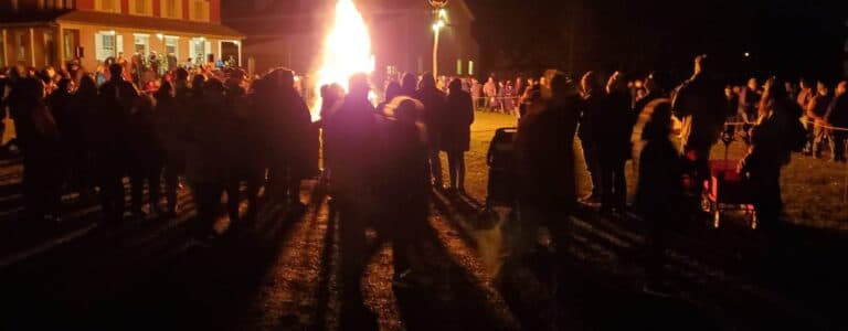 A large crowd gathers around a bonfire at night, with a building visible in the background.