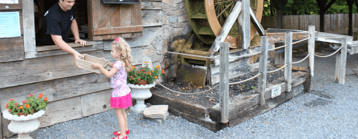 A child in a pink outfit receives a box from a person at a wooden stand next to a water wheel.