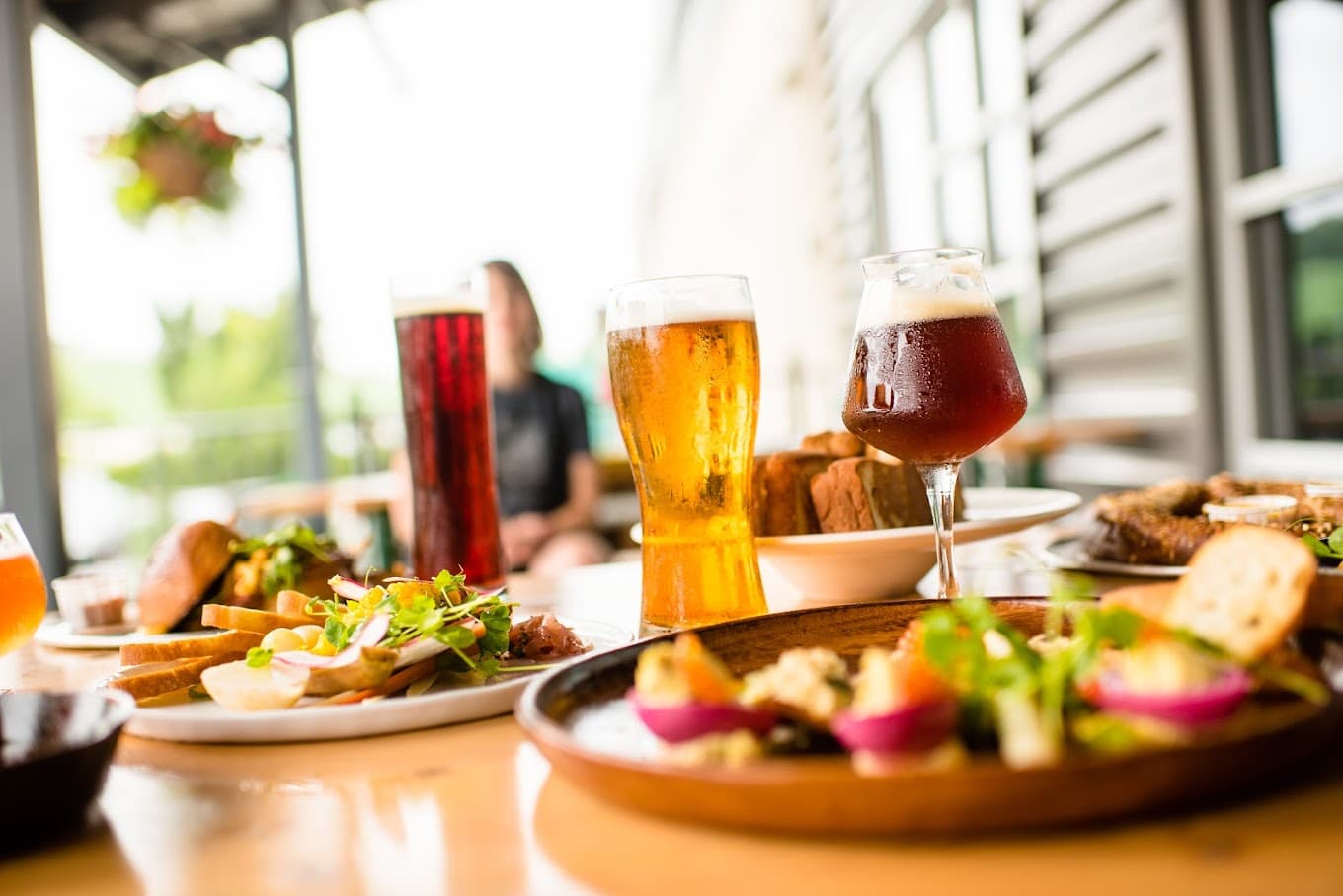 A table filled with various dishes and three glasses of beer in a bright outdoor setting.