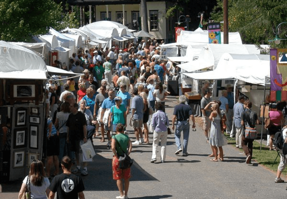 A large crowd explores an outdoor market with white tents and various displays.