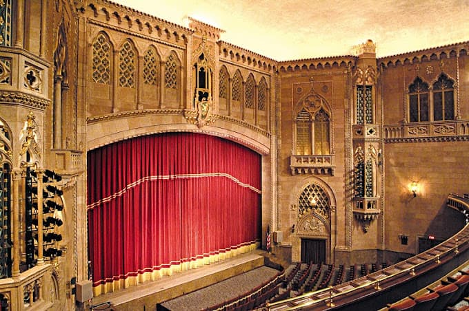 An ornate theater interior featuring a crimson curtain and intricate architectural details.