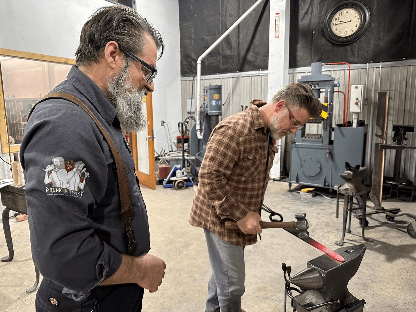 Two men observe and work with heated metal at an anvil in a workshop.