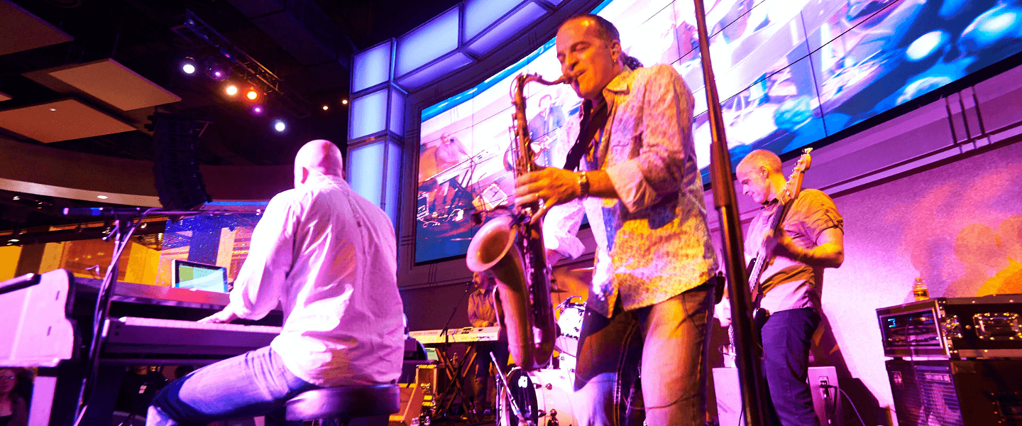 A live band performs on stage with a saxophonist in the foreground and vibrant lighting in a lively atmosphere.