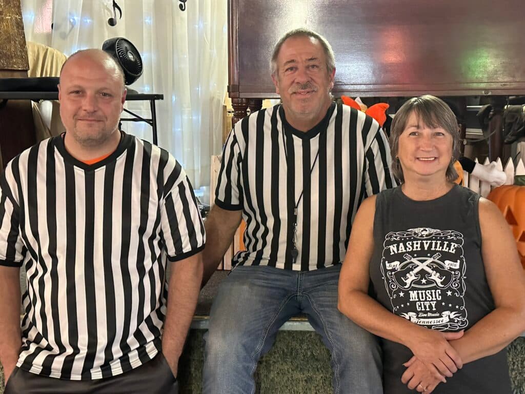 Three individuals wearing black and white striped referee shirts pose together in a casual setting.