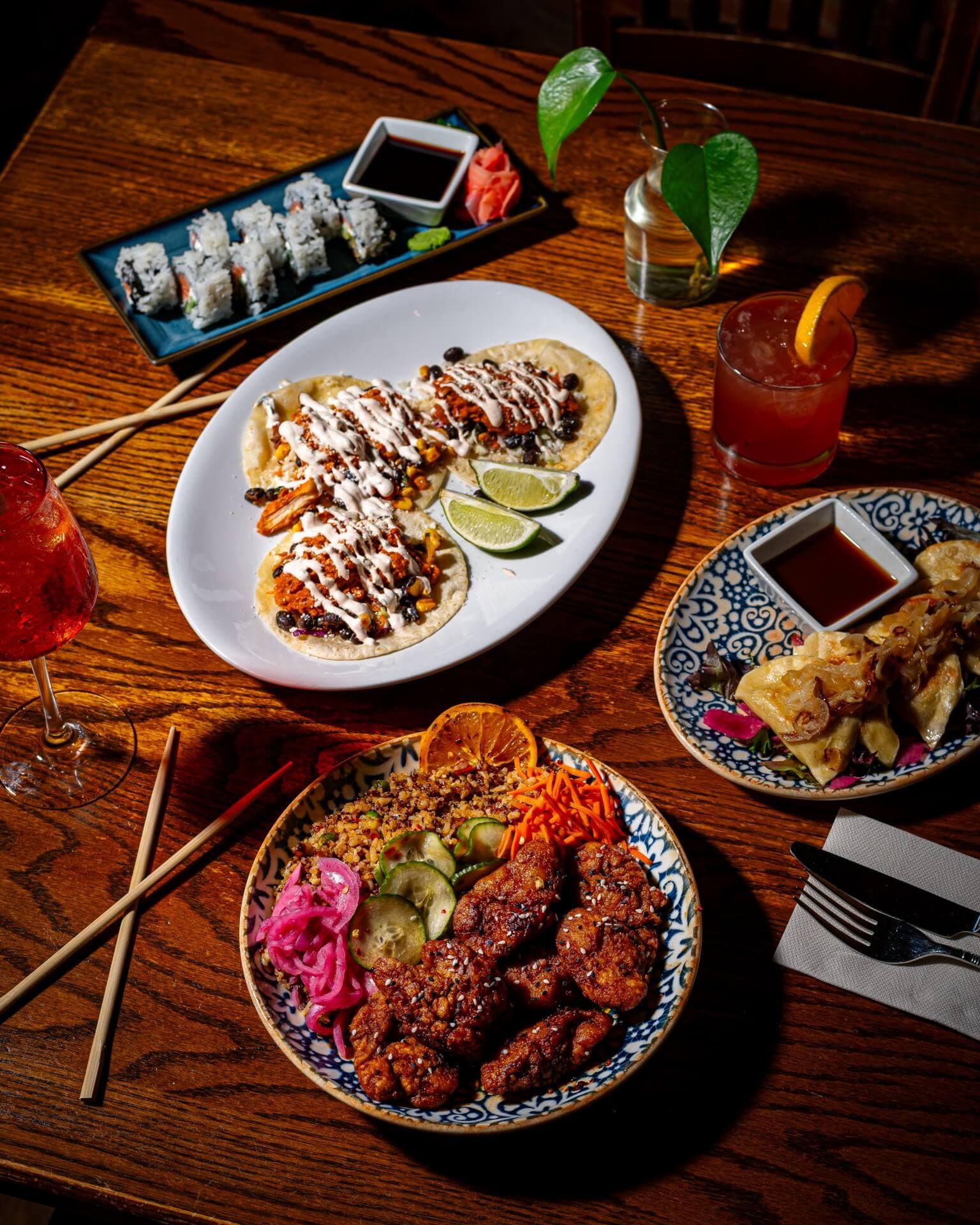 A colorful spread of tacos, chicken, sushi, and drinks arranged on a wooden table.